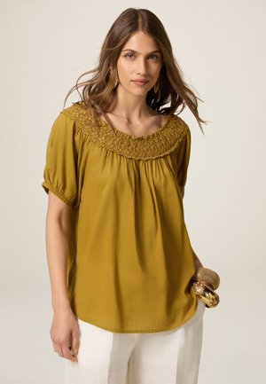 Blusa amarilla mostaza con cuello fruncido y cortas mangas abullonadas, con tejido suave y una forma holgada y fluida.