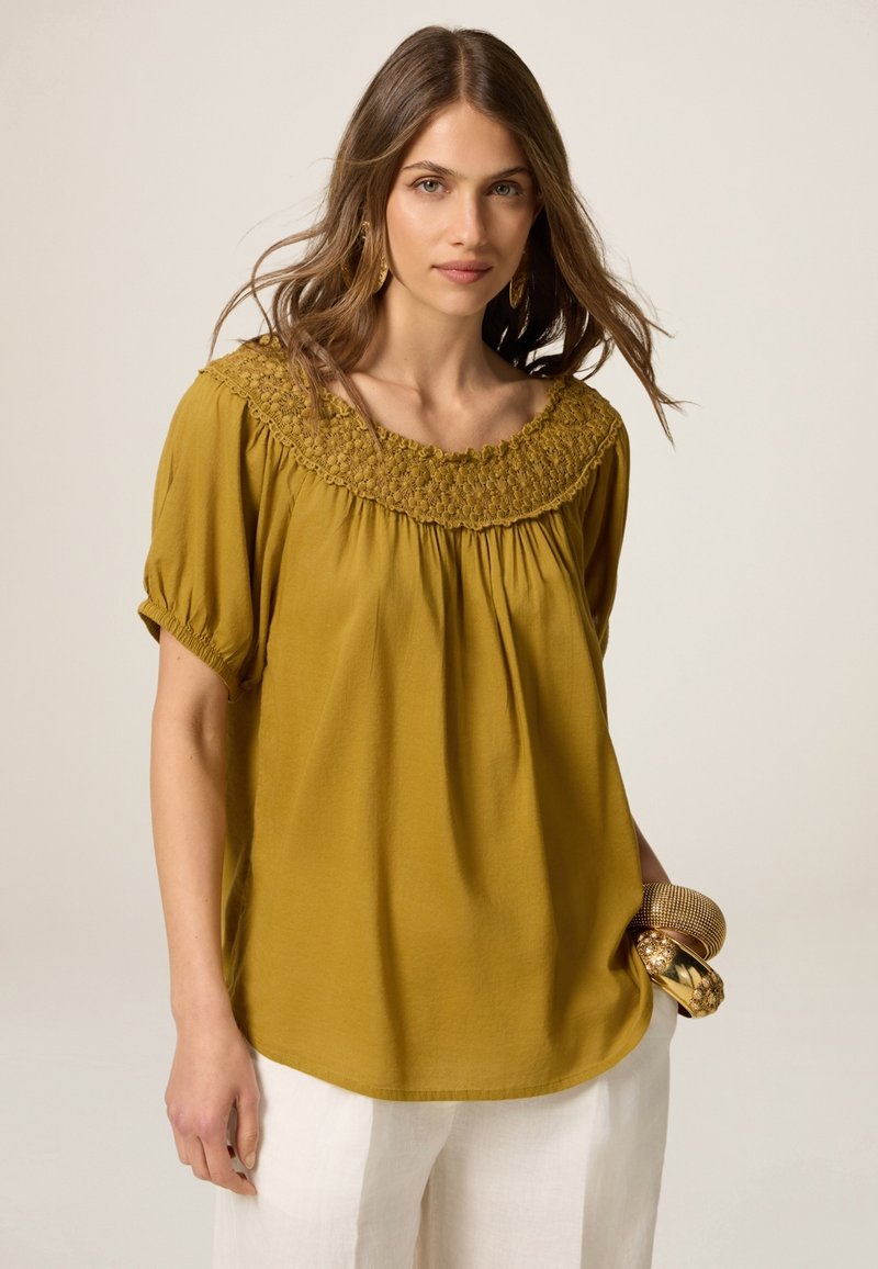 Mosterdgele blouse met een smockhals en korte pofmouwen, gemaakt van zachte stof en met een losse, vloeiende vorm.