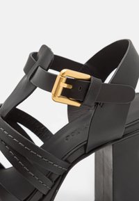 Black leather heeled sandal with a wide strap design, featuring a gold metal buckle, gray stitch details, and a chunky black platform heel.