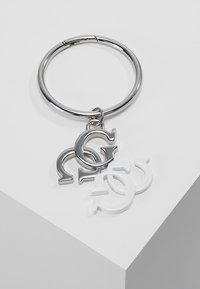 Sleutelhanger met een zilveren metalen ring en drie bevestigde bedels: twee in witte acryl en één in gepolijst zilver, met gestileerde ontwerpen van de letter "G".