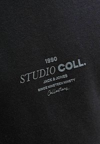 Tela negra con un acabado texturizado presenta un logo impreso en gris: "STUDIO COLL. 1990 JACK & JONES DESDE MIL NOVECIENTOS NOVENTA Colecciones."
