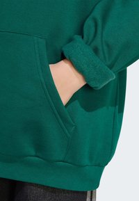 Felpa verde con cappuccio e texture morbida, caratterizzata da una tasca frontale e polsini arrotolati. Il tessuto appare spesso e accogliente, adatto per il clima fresco.