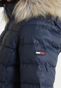 Tmavě modrá prošívaná bunda s kapucí lemovanou kožešinou, prošívaným vzorem a malou nášivkou loga v červené, bílé a modré na rukávu.