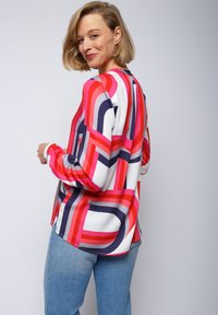 Langärmlige Bluse in mehreren Farben mit einem auffälligen geometrischen Muster in Rot, Pink und Marineblau auf einem weißen Hintergrund; leichtes Material, lockere Passform.