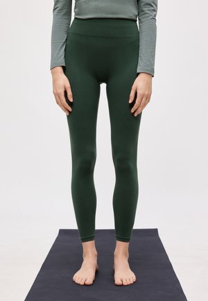 Persona de pie descalza sobre una esterilla de yoga oscura, vestida con leggings ajustados verde oscuro y una camiseta gris de manga larga, corta a la mitad del torso.
