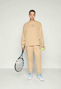 Bežan sweatshirt in ujemajoči se trenirki z modro logotipno detajlijo, drži teniško lopar in žogico. Bombažen material, sproščen kroj, preprost dizajn.