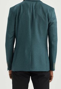 Blazer teal con tessuto testurizzato, rever a punta e tre bottoni su ciascun polsino. Vestibilità sartoriale, spacco singolo sul retro, abbinato a pantaloni neri.