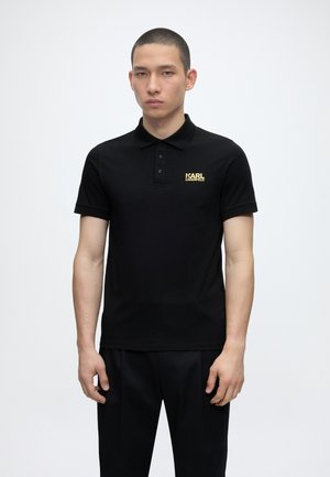 Fiatal férfi áll, fekete galléros pólót visel arany "KARL LAGERFELD" logóval a bal mellrészen, egyszínű fehér háttér előtt.