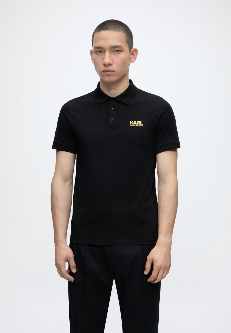 Νεαρός άνδρας στέκεται φορώντας μαύρο πόλο με χρυσλό λογότυπο "KARL LAGERFELD" στο αριστερό στήθος, σε απλό λευκό φόντο.