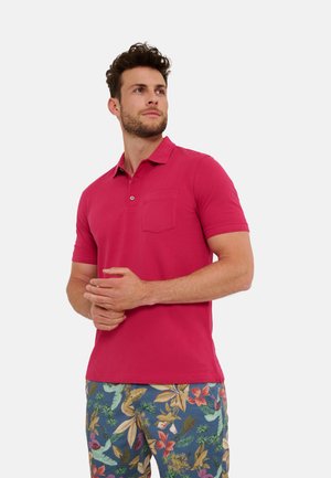 Enganliegendes pinkes Poloshirt mit Kragen und einer Tasche. Getragen mit gemusterten blauen Shorts, die bunte Blätter und Blumen zeigen.