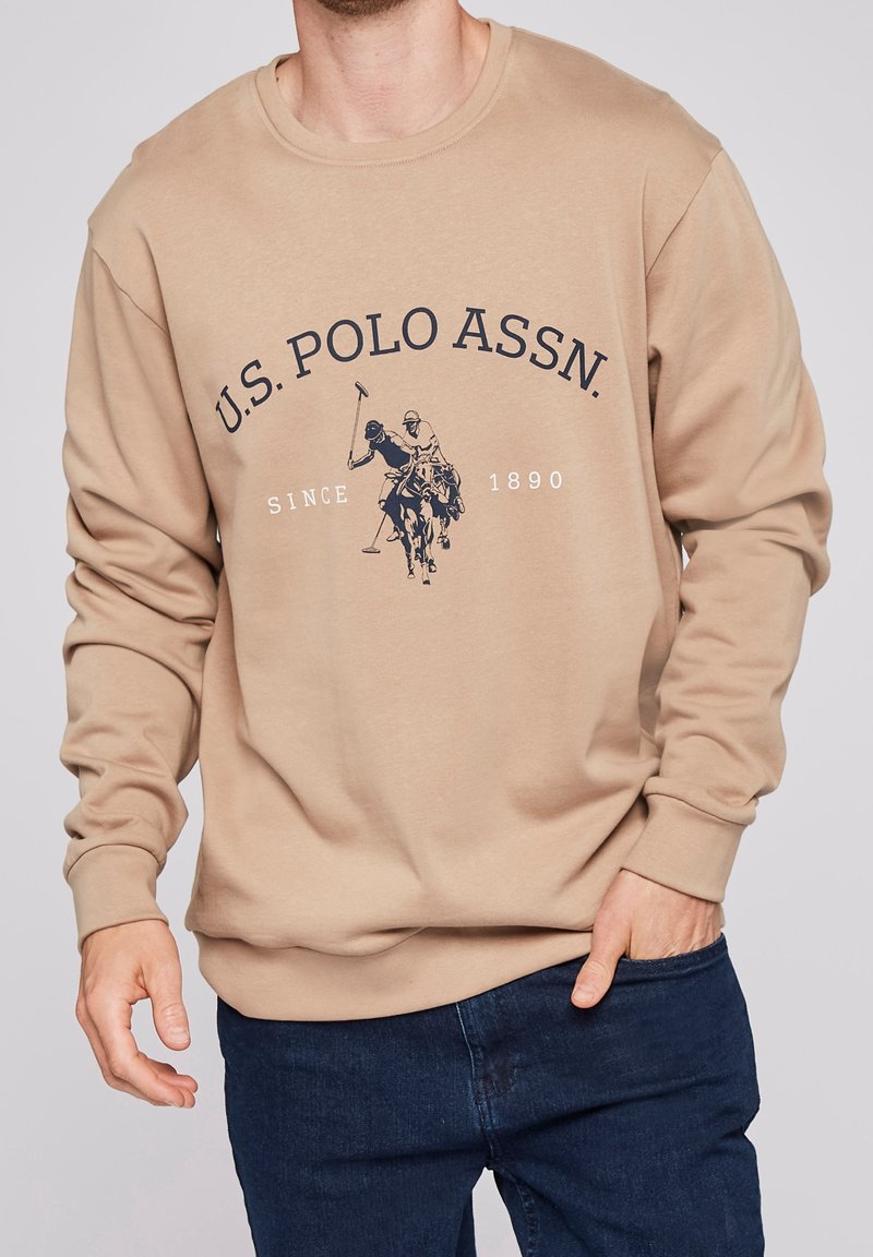 Beige sweatshirt i mjukt tyg, med långa ärmar och rundad halsringning. En mörk grafik av polo-spelare och text är centrerad på framsidan.