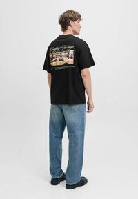 Sort bomulds T-shirt med en ryggrafik, der viser et fotografi og tekst; parret med lyseblå denimjeans og sort fodtøj.