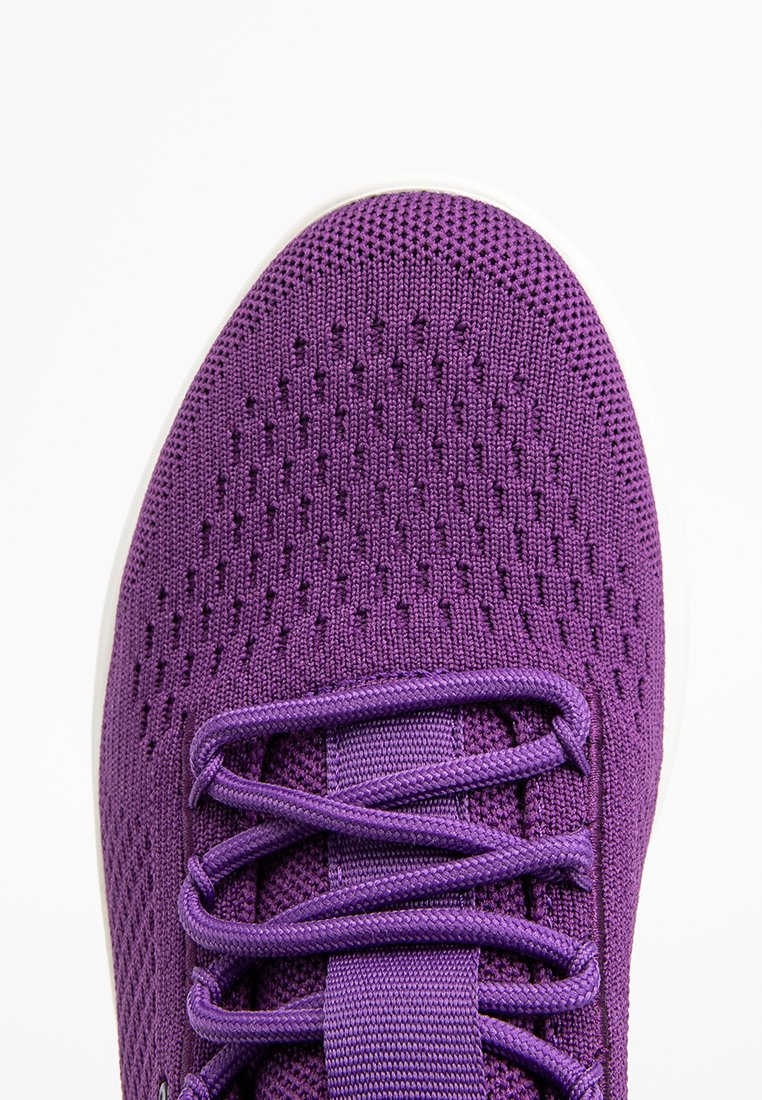 Primo piano di una sneaker lavorata a maglia viola con lacci incrociati su uno sfondo bianco, evidenziando la texture e il materiale traspirante.