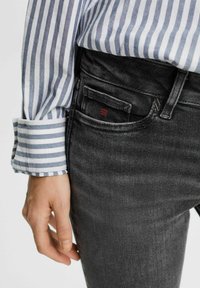 Personne portant un jean noir et une chemise à manches longues rayée bleu et blanc, main détendue sur le côté près de la poche avant.