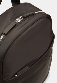Mochila negra con tejido suave, que cuenta con un bolsillo delantero con cremallera y herrajes en negro mate. Forma redondeada con un brillo sutil.