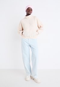 Helles beige Fleece-Pullover mit einem Halb-Reißverschluss an der Vorderseite, gerippten Bündchen und einer Kängurutasche, kombiniert mit hellblauen Jeans und creamfarbenen Sneakers.