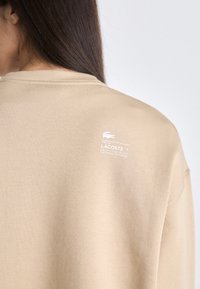 Sudadera de color beige claro con cuello redondo y mangas largas. Presenta un pequeño logotipo blanco y texto impreso en la parte superior de la espalda. Textura suave.