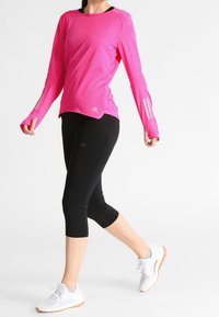 Kobieta idąca, ubrana w jasnoróżową, długą bluzę sportową, czarne legginsy do kostek oraz białe buty do biegania z gumową podeszwą.