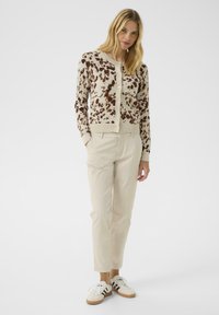 Cream cardigan with brown spotted pattern, featuring a button front and ribbed cuffs. Paired with light beige pants and white sneakers.