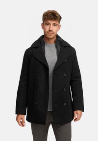 Schwarzer Wollpeacoat mit doppelt-knöpfbaren Knöpfen, breiten Revers und leicht gepolstertem Kragen, getragen über einem hellgrauen Pullover.