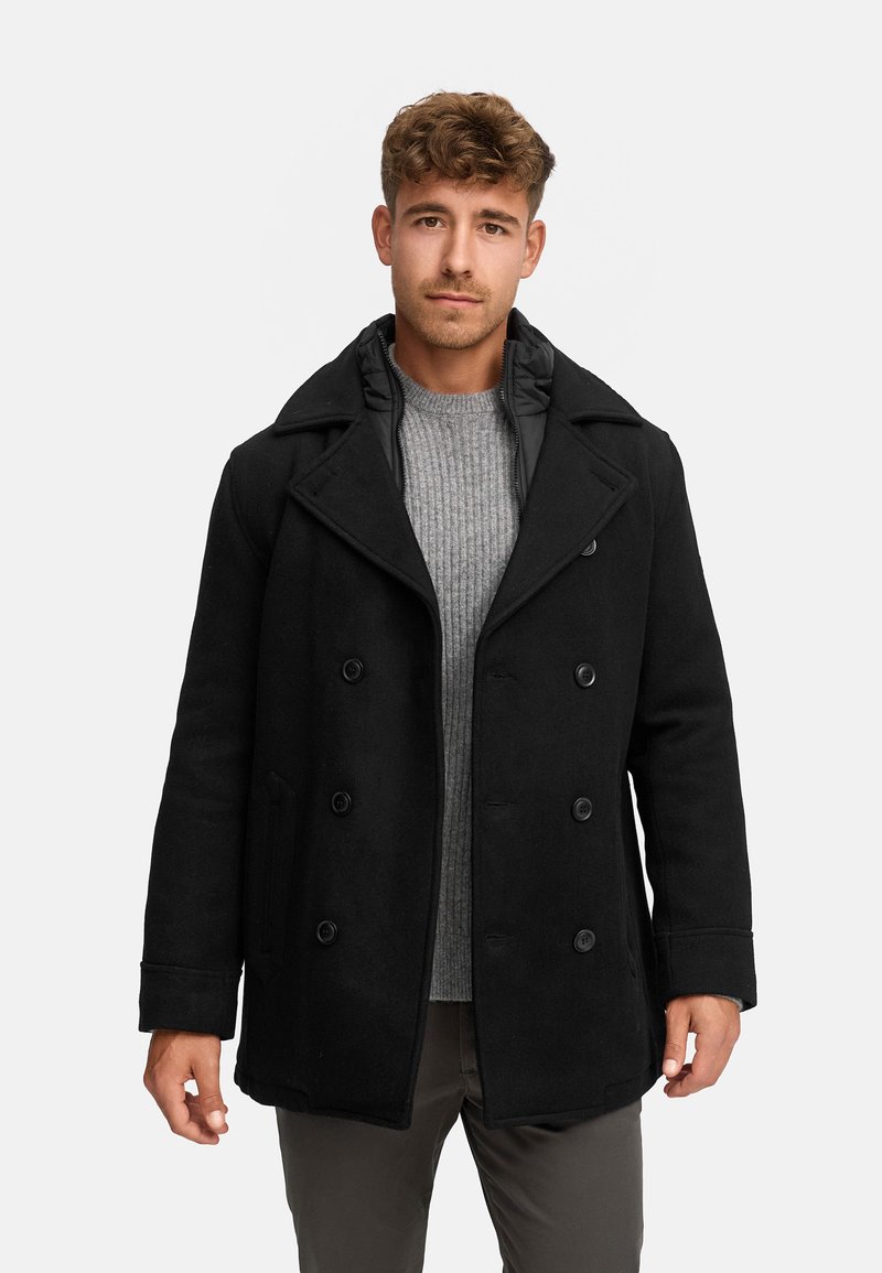Schwarzer Wollpeacoat mit doppelt-knöpfbaren Knöpfen, breiten Revers und leicht gepolstertem Kragen, getragen über einem hellgrauen Pullover.