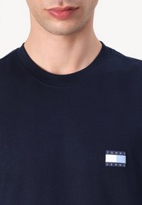 Navy blue cotton t-shirt with short sleeves, round neckline, and a small branded patch featuring a blue and white logo on the chest.