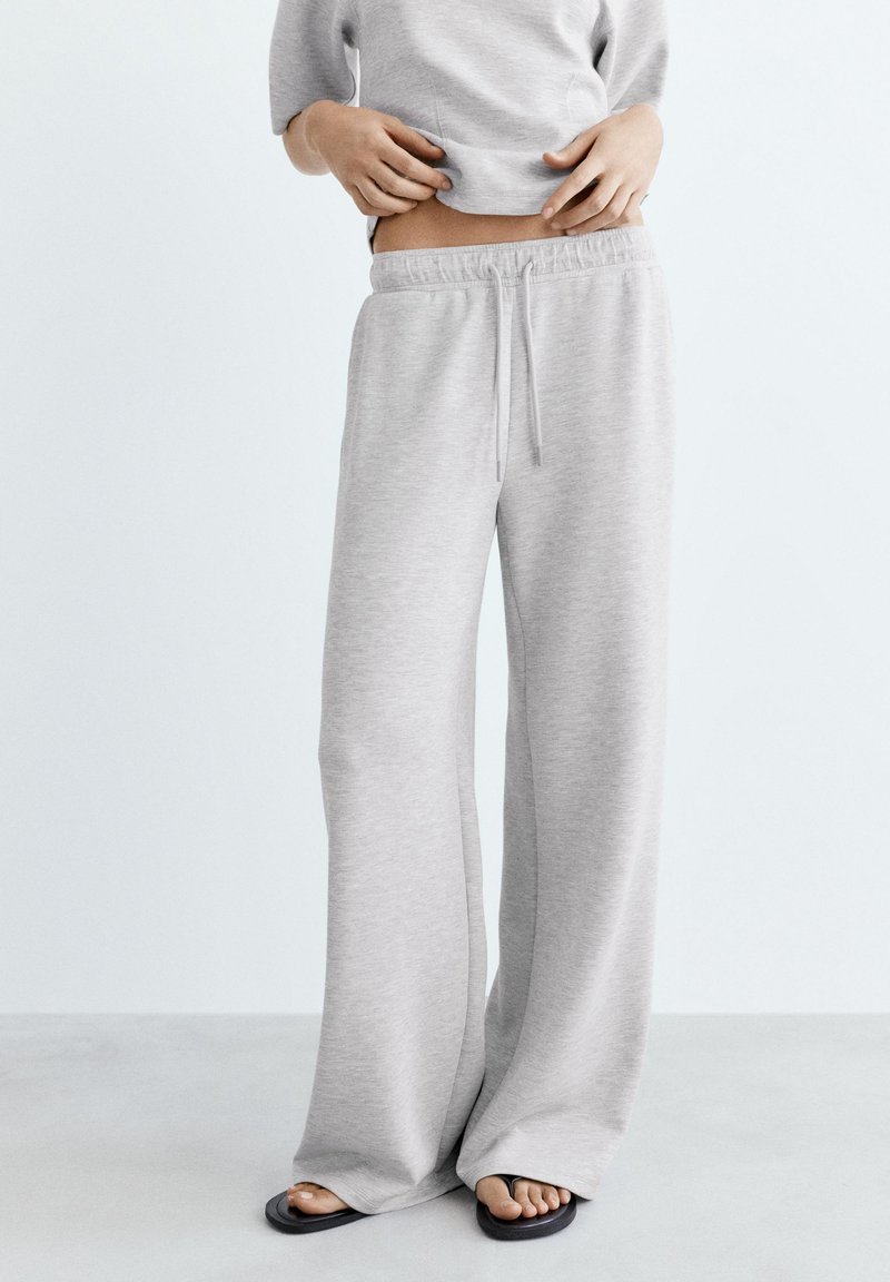 Personne portant un pantalon de survêtement gris clair à jambes larges et des tongs noires, soulevant un sweat-shirt gris pour dévoiler le ventre nu.