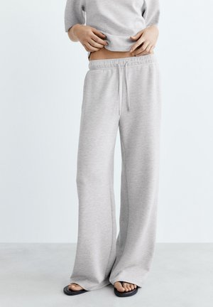 Persona che indossa pantaloni della tuta larghi grigio chiaro e infradito neri, sollevando una felpa grigia per mostrare l'addome scoperto.