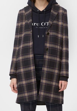 Frau trägt einen dunkel karierten langen Mantel über einem marineblauen Hoodie mit weißem Schriftzug und passenden dunkel karierten Hosen.