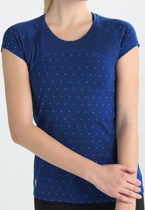 Kvinde iført en mørkeblå kortærmet skjorte med små hvide prikker, stående med armene krydset ved livet.