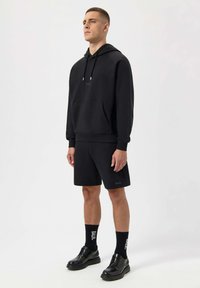 Black hoodie with a front pocket and drawstring, paired with black shorts. Black socks and shiny black dress shoes complete the outfit.