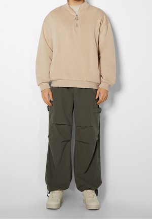 Pull-over beige à demi-fermeture éclair associé à un pantalon cargo vert foncé et des baskets beiges, porté par une personne debout sur un fond blanc.