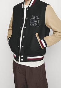 Zwarte en tan varsityjack met drukknoopsluiting, geribde manchetten en een geborduurd logo. Beschikt over twee zijzakken en een contrasterende gestreepte zoom.