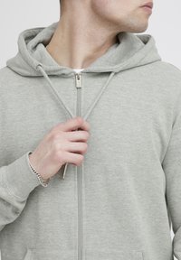 Felpa grigia chiara con zip, cappuccio regolabile, tasche frontali e tessuto testurizzato. Presenta una zip argentata e un design minimalista.