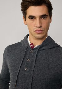 Felpa grigia lavorata a maglia con cappuccio e coulisse, con tre bottoni scuri e una camicia bordeaux in contrasto sotto. Texture del tessuto liscia.