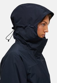 Giacca impermeabile blu con colletto alto e cappuccio regolabile, caratterizzata da una texture liscia e un design minimalista.