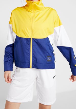 Personne portant une veste zippée jaune, blanche et bleue avec un écusson logo et un short de sport blanc avec le logo Nike noir sur fond uni.