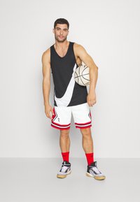 Camiseta de baloncesto sin mangas en blanco y negro, pantalones cortos atléticos blancos con acentos rojos, calcetines rojos y zapatillas negras, sosteniendo un balón de baloncesto.