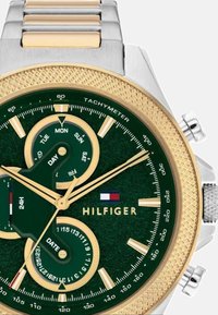 Edelstahlarmbanduhr mit goldenen Akzenten, grünem strukturiertem Zifferblatt, Hilfszifferblättern für Datum und Wochentag sowie Tachymeter. Mit Markenlogo.