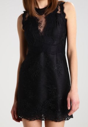 Mouwloze zwarte kanten mini-jurk met doorzichtige bloemkanten halslijn en geschulpte zoom, gedragen door een staand persoon met schouderlang bruin haar.