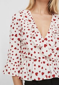 Blusa bianca con motivo a cuori rossi, scollo a V profondo, bordi arricciati e orlo a peplum. Maniche lunghe con polsini svasati. Tessuto dalla trama liscia.