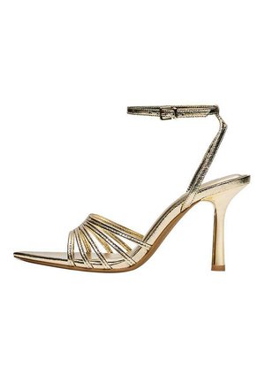 Kultainen remmillä varustettu korkeakorkoinen sandaali nilkkaremmi, avoin varvas ja ohut piikkikorko valkoisella taustalla.