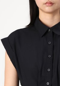 Camisa preta de colarinho com mangas curtas e dobradas, apresentando uma frente com botões e um tecido ligeiramente texturizado.