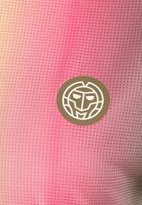 Tejido texturizado en degradado rosa, con un logo redondo que presenta un rostro estilizado en blanco sobre un fondo marrón.