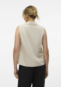 Top sans manches beige avec un col et une texture douce. L'arrière présente un ourlet incurvé et une finition lisse. Assorti avec un pantalon noir.