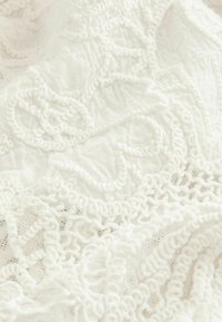 Tissu blanc avec des détails en dentelle complexe, présentant des motifs texturés et des bords froncés. Le matériau semble doux et léger.