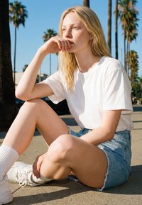 Ung kvinde med langt blondt hår, hvid t-shirt, denimshorts og sneakers, der sidder på fortovet nær palmer på en solrig dag.