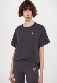 Rövid ujjú, laza szabású, sötétszürke póló kerek nyakkal és diszkrét fémes logóval a mellkason, párosítva illő leggingsel.