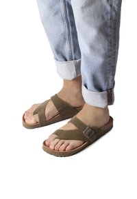 Brown suede sandals with a cross-strap design, featuring a buckle accent and textured rubber sole, worn with cuffed light denim.