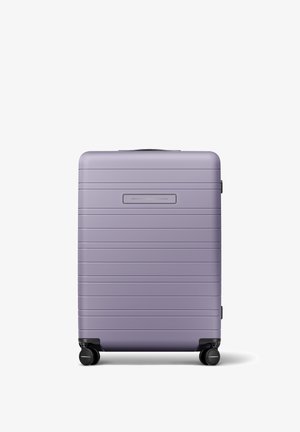 Valise rigide de couleur violet clair, avec une texture nervurée et une poignée supérieure, équipée de quatre roues noires pour une mobilité facile.
